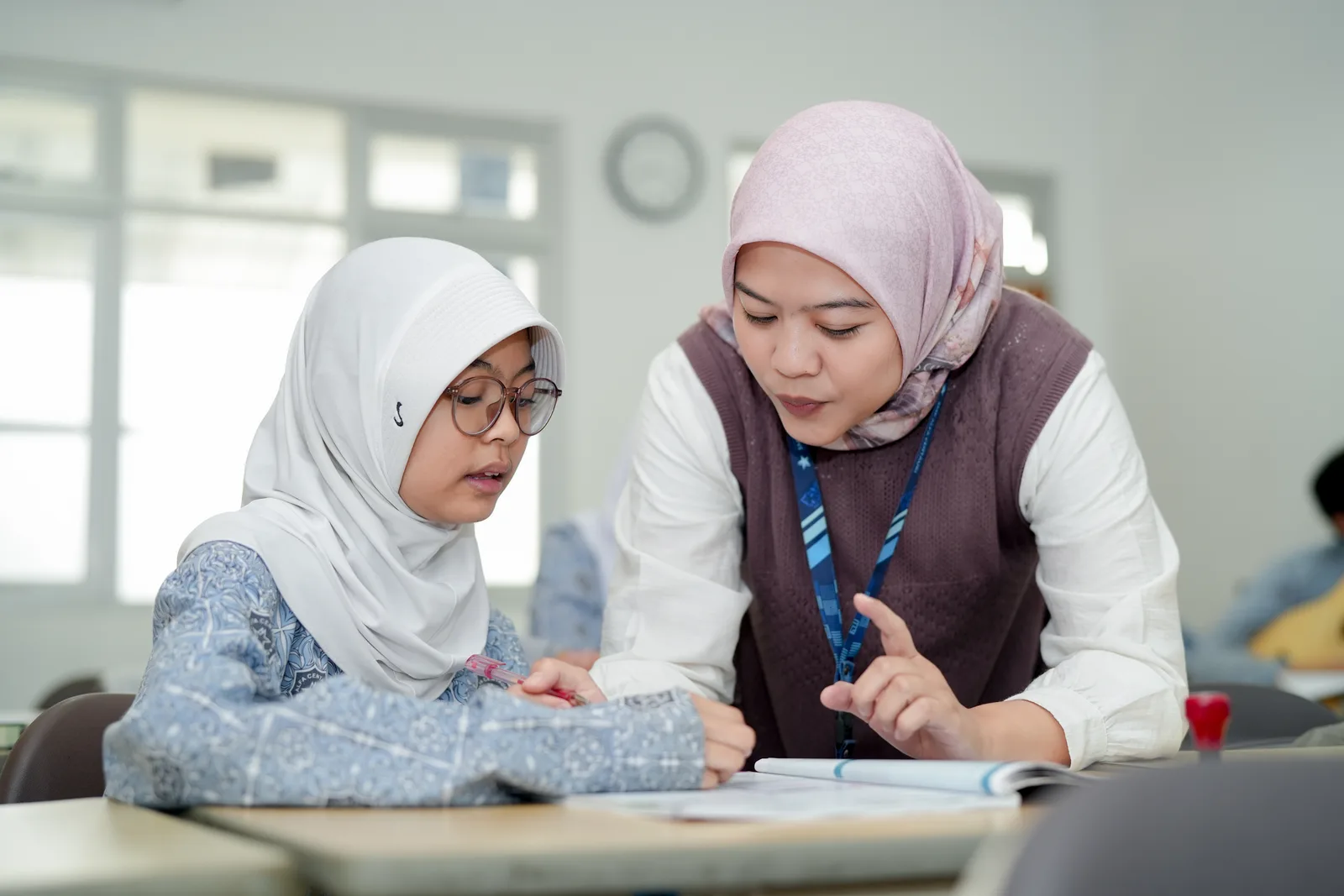 Guru pendamping membimbing siswi belajar secara personal di ruang kelas.